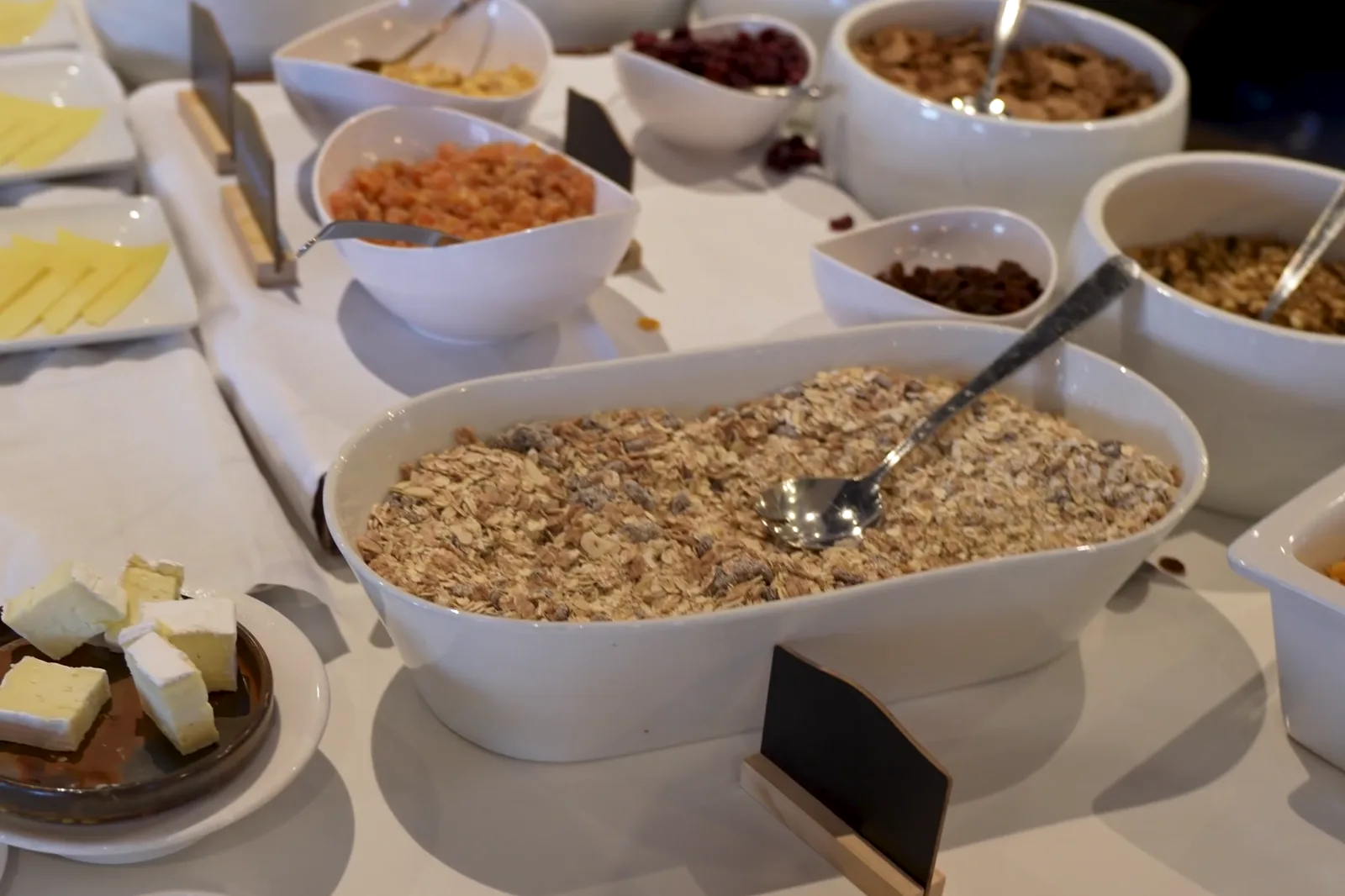 Self-service line with oat flakes, granola, dried fruit and cheese in china bowls, with blank slate labels in front.