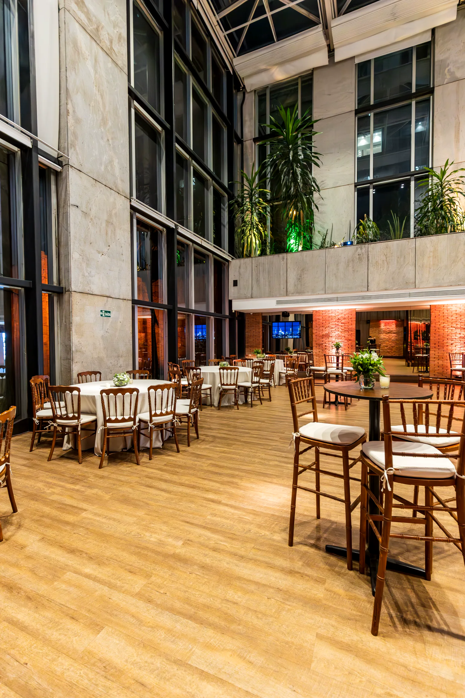 Salón de hotel de doble altura con mesas vestidas y vegetación, listo para recibir.