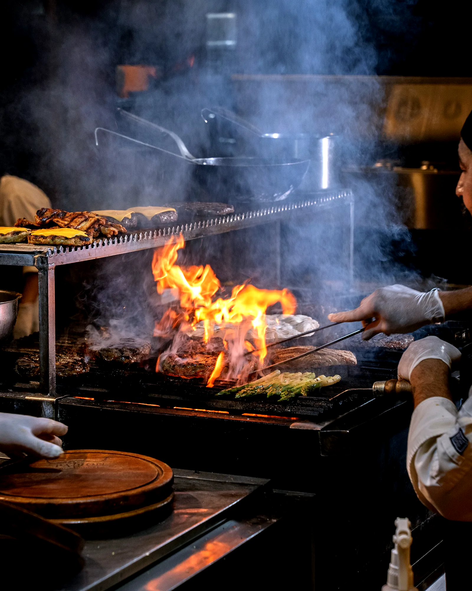 Estación de parrilla en vivo durante un servicio, con la llama alta bajo las piezas y el cocinero volteándolas con pinzas.