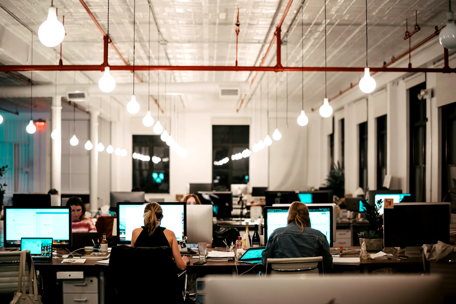 Oficina de planta abierta al final de la tarde, con los puestos ocupados bajo las lámparas encendidas.