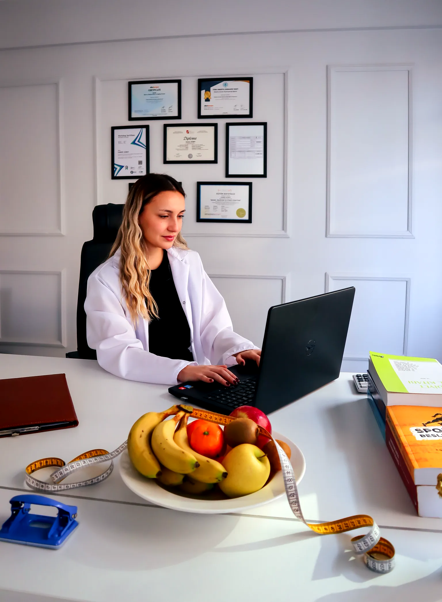 Consulta de nutrición: profesional en bata revisando un plan alimentario, con fruta fresca y cinta métrica sobre el escritorio.
