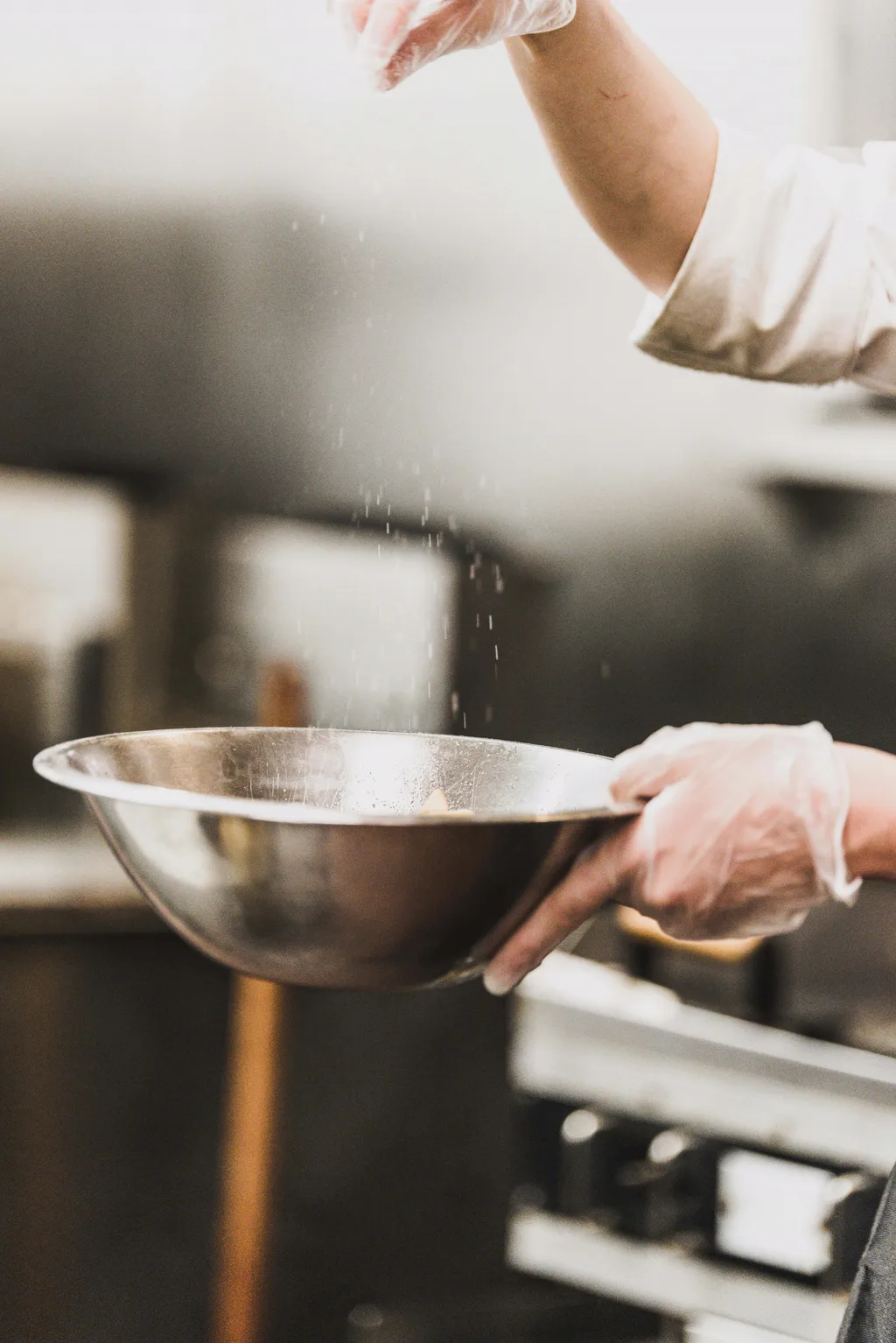 Manos con guante sazonando sobre un bol de acero inoxidable en la cocina de producción.