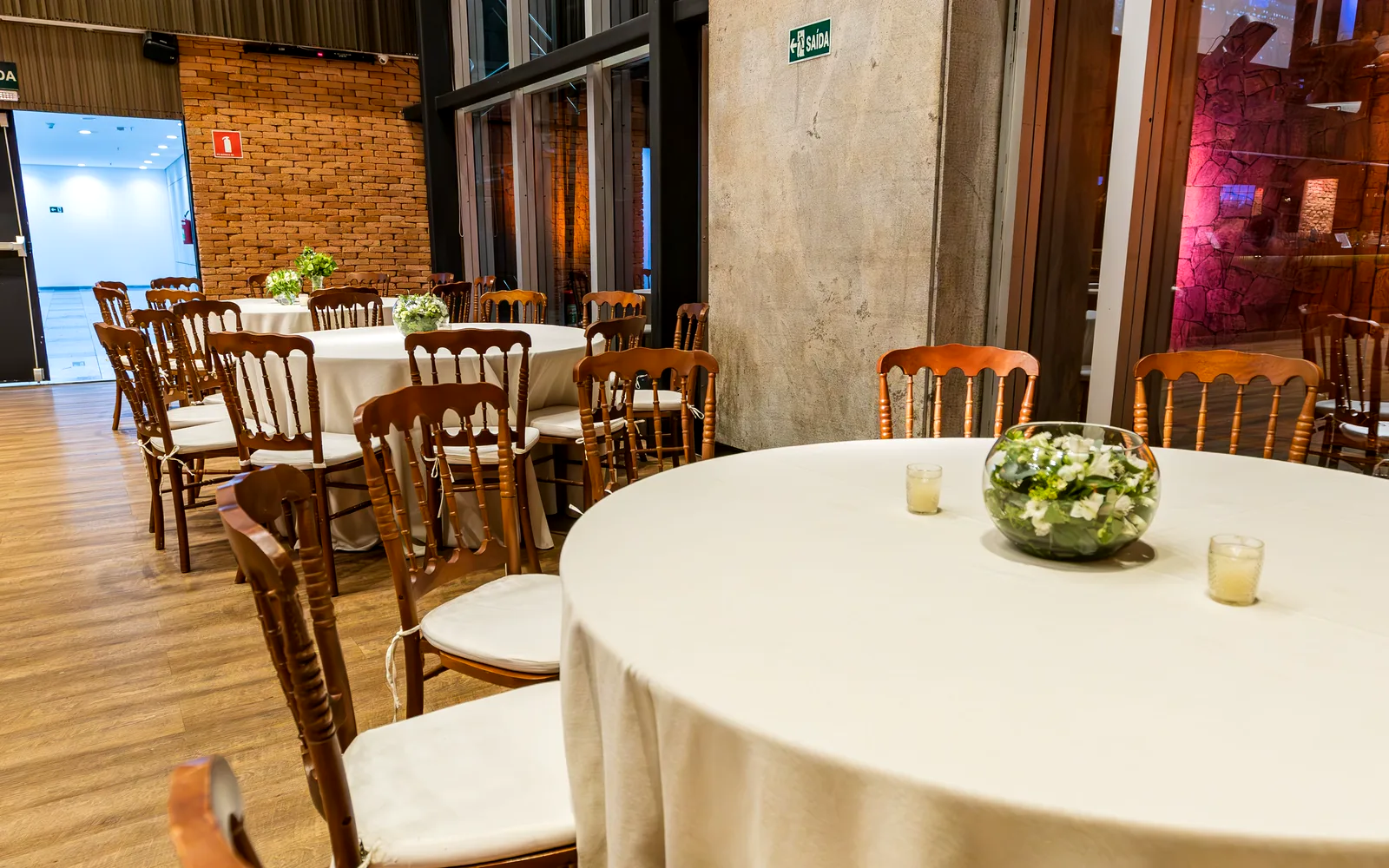 Salón montado para un banquete corporativo: mesas redondas con mantel, sillas de madera y un centro floral bajo la luz de los ventanales.