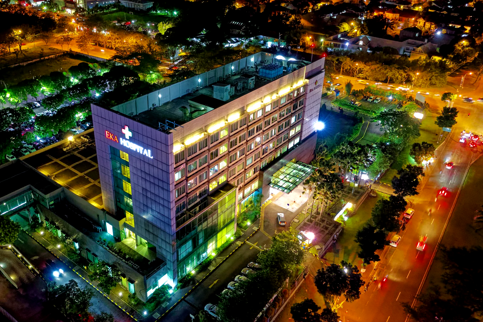 Vista aérea nocturna de un hospital iluminado, con la avenida de acceso en movimiento.